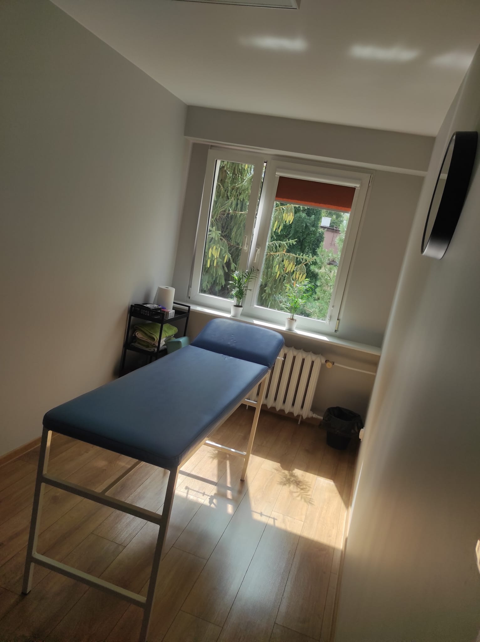 Gabinet zabiegowy w zakładzie fizjoterapeutycznym w Płocku z niebieskim stołem do masażu, dużym oknem z widokiem na zieleń i stonowaną, relaksującą atmosferą. Gabinet zabiegowy w zakładzie fizjoterapeutycznym w Płocku z niebieskim stołem do masażu, dużym oknem z widokiem na zieleń i stonowaną, relaksującą atmosferą.